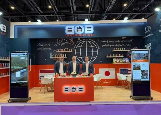 Three staff behind the orange counter of the BOB stand, with two screens and flags of the countries of origin.