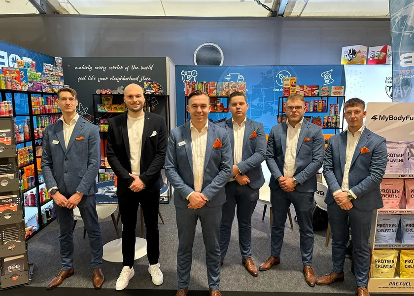 Six BOB Distribution staff in suits in front of their trade fair stand with shelves full of sweets and soft drinks.