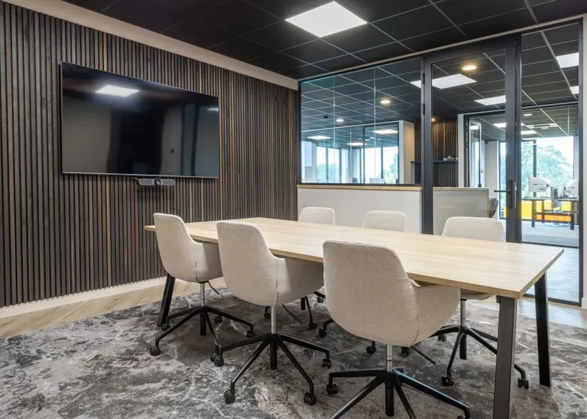 A boardroom with a long table, beige chairs, a wooden wall and a large screen.